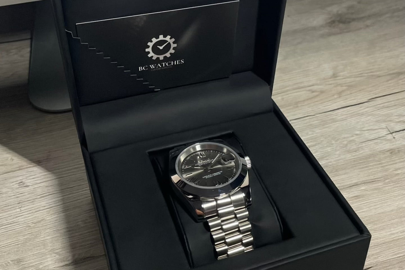 Silver watch in a black box with 'BC Watches' branding on a wooden surface
