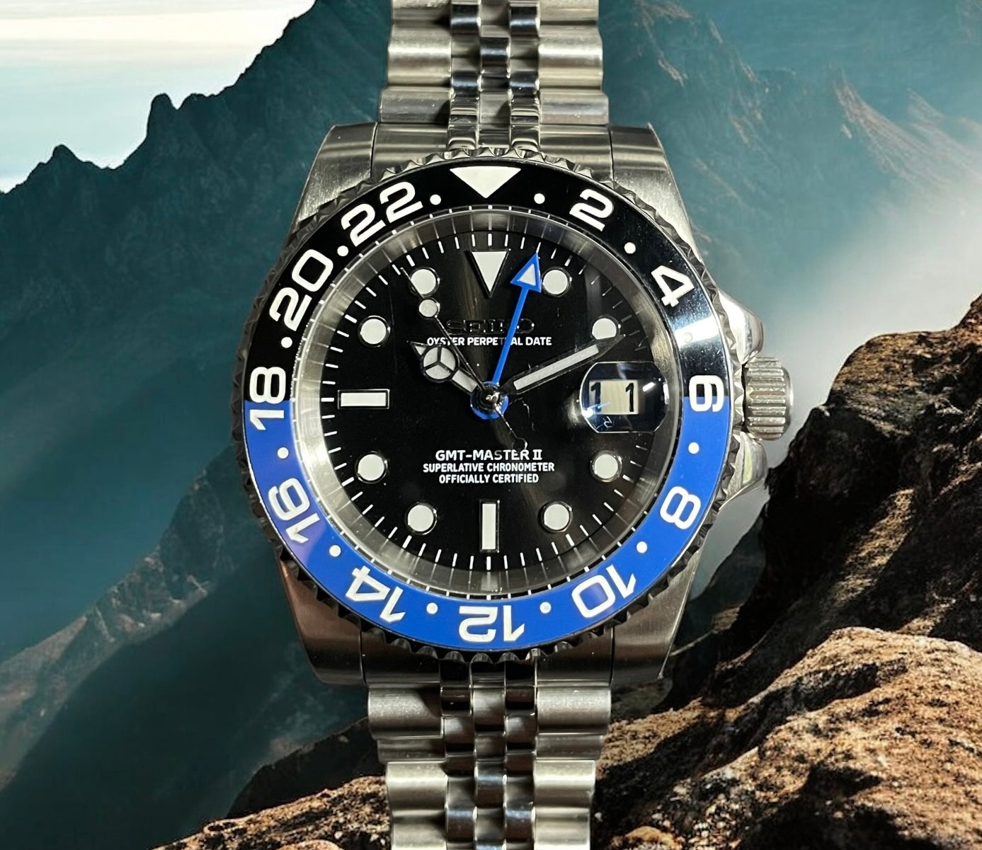Silver watch with black and blue bezel on a rocky surface with mountain background